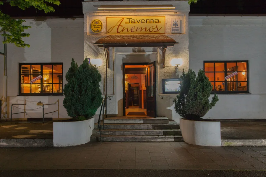 Fassade der Taverna Anemos bei Nacht mit Leuchtschild über dem Eingang und Buchsbaum in Kübeln